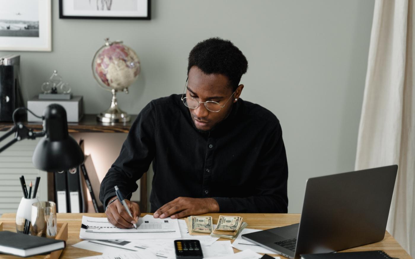 A young man sitting at a desk. There is a calculator and money on the desk, and the man is writing on a chart.